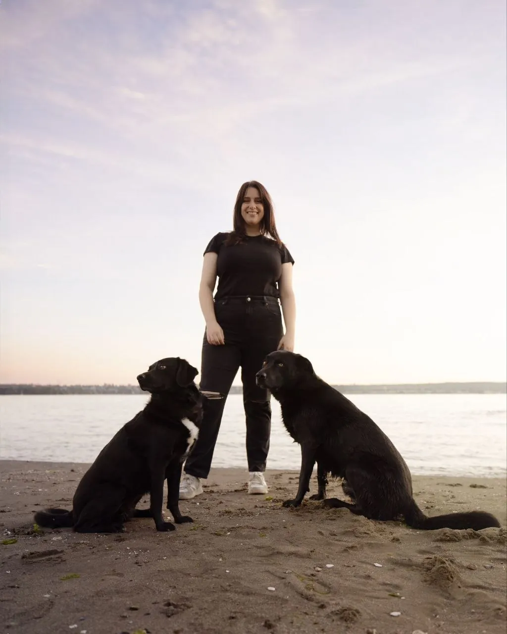 Two calm black Labradors sitting with Lea on the beach at sunset